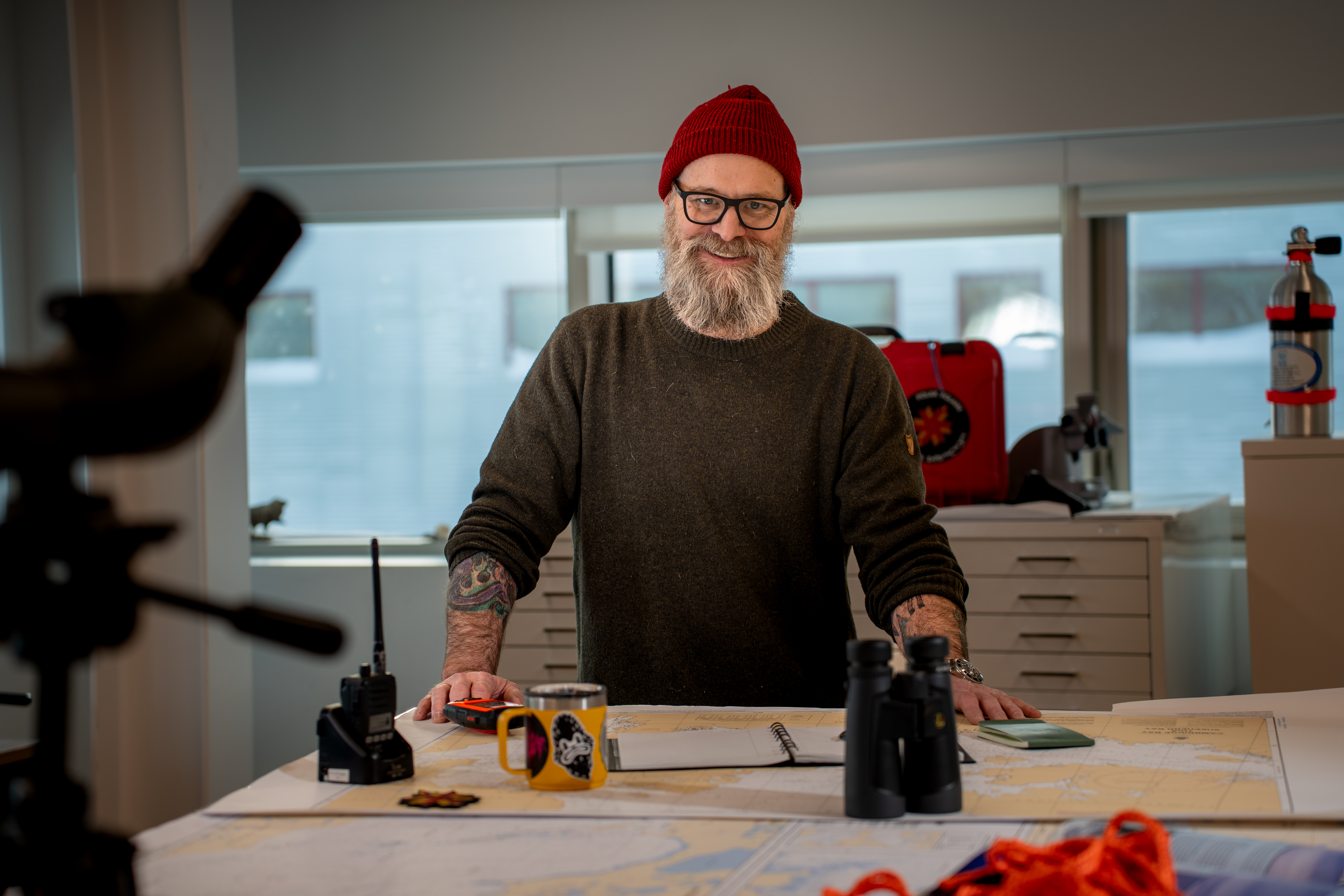 Man with beard and hat on, stands behind a table with hands resting on tabletop.
